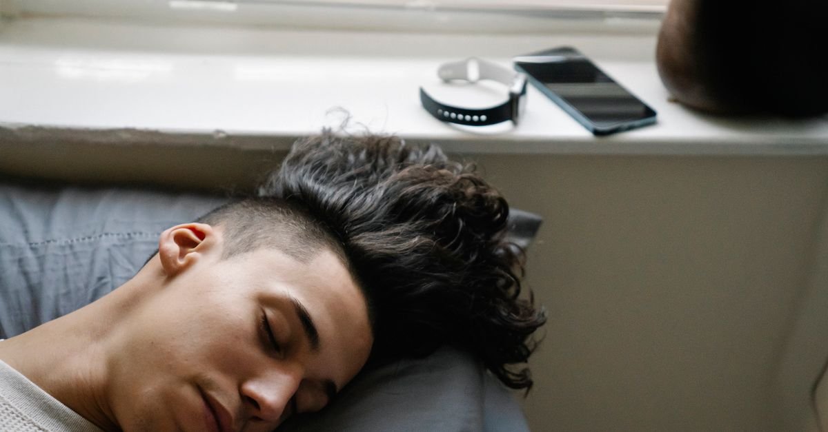 High angle of young Hispanic male with curly hair lying on bed near windowsill and sleeping in morning at home