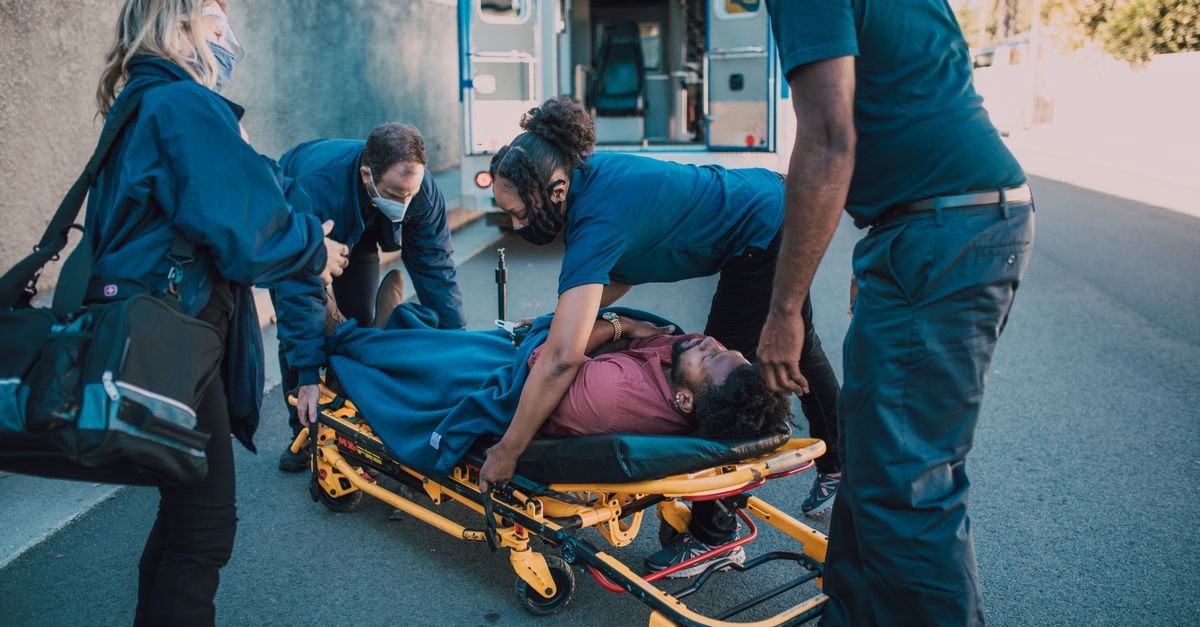 Paramedics carefully assist a patient onto a stretcher near an ambulance. Outdoor medical emergency scene.