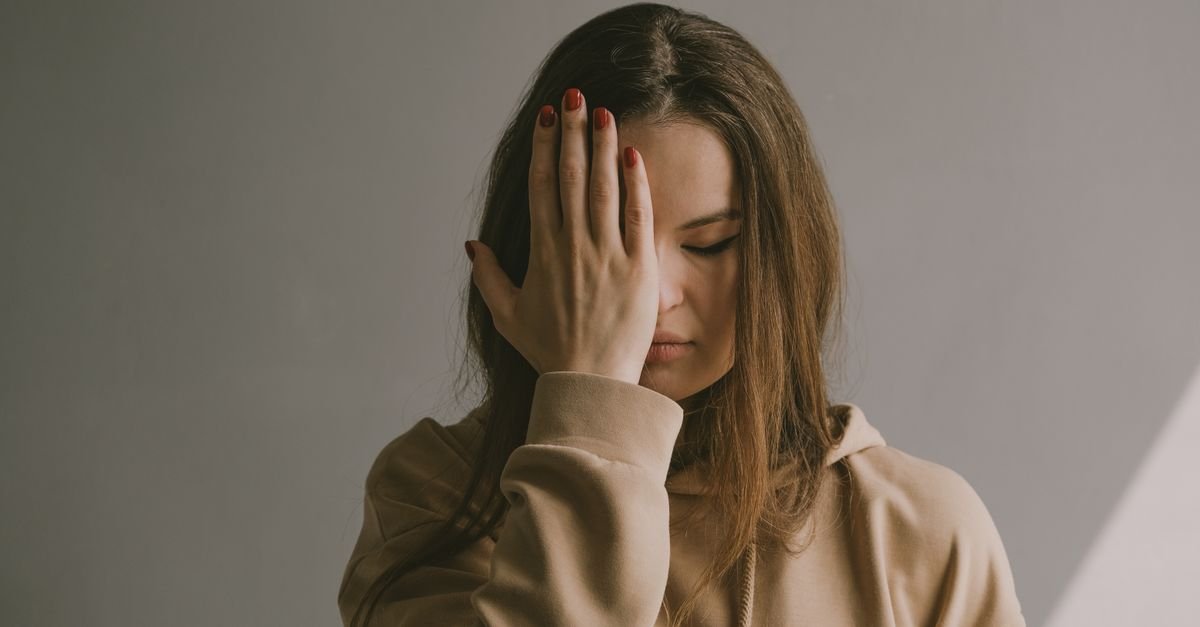 A woman with brown hair wearing a hoodie covers her face with her hand, depicting emotion.