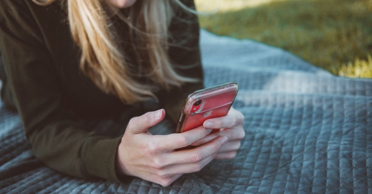 Young woman lying on a blanket outdoors using a smartphone on a sunny day.
