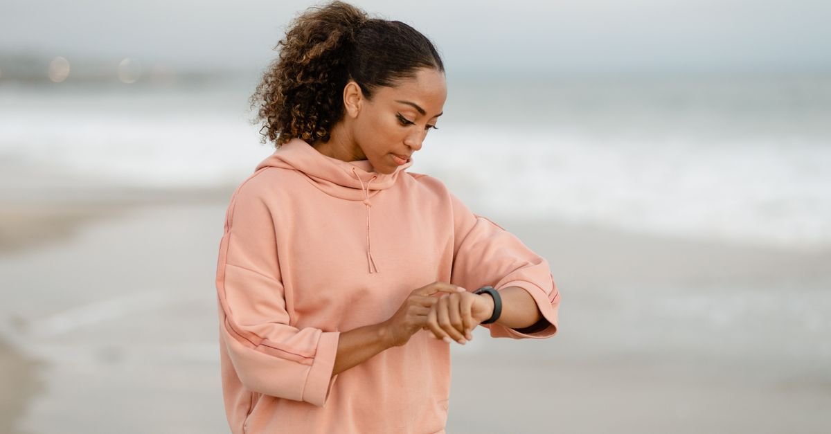 A woman in casual attire checks her smartwatch while standing on a sandy beach.