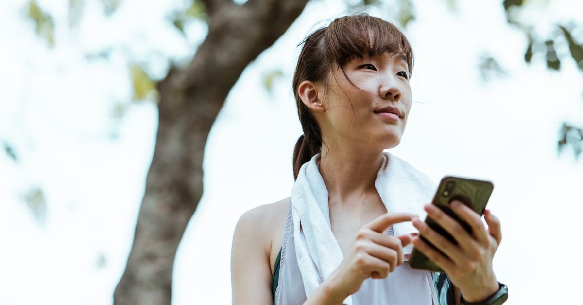 From below of content Asian sportive female with towel on shoulders and smart watch chatting on cellphone and looking away in park