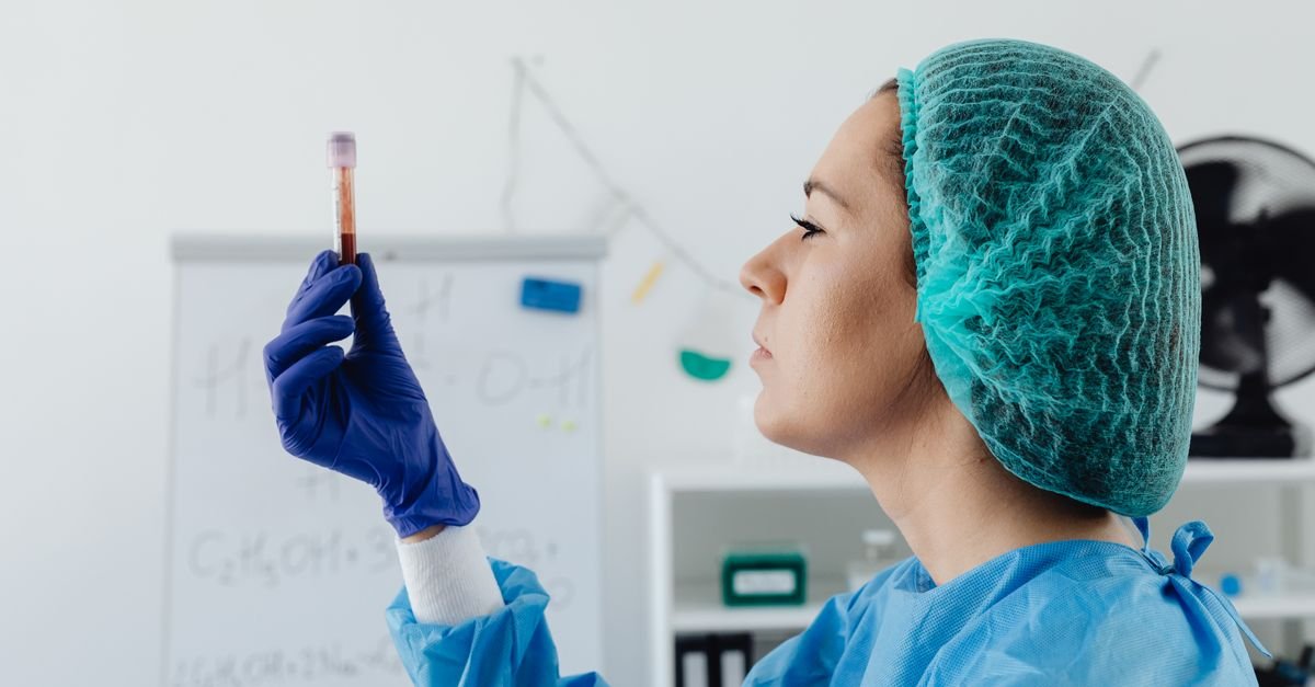 A female scientist in protective gear inspects a blood sample vial in a lab setting.