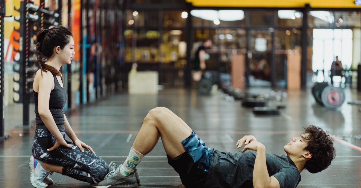 Asian couple engaged in workout session performing sit-ups in a well-equipped gym setting.