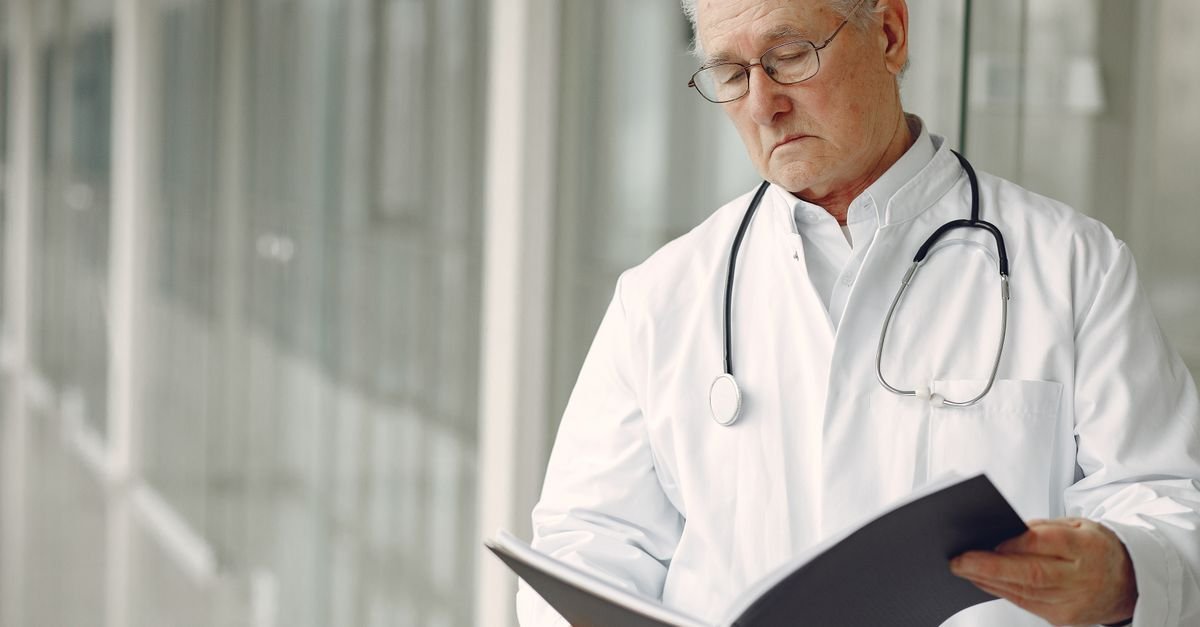 Crop concentrated professional senior male physician in white robe with stethoscope standing in modern hospital hallway and reading medical case records in daylight