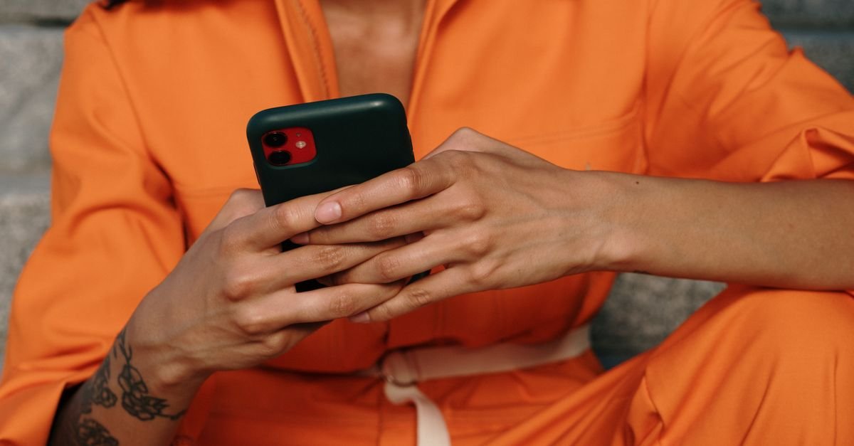 A person in an orange outfit using a smartphone, showcasing a close-up of hands and tattoo.