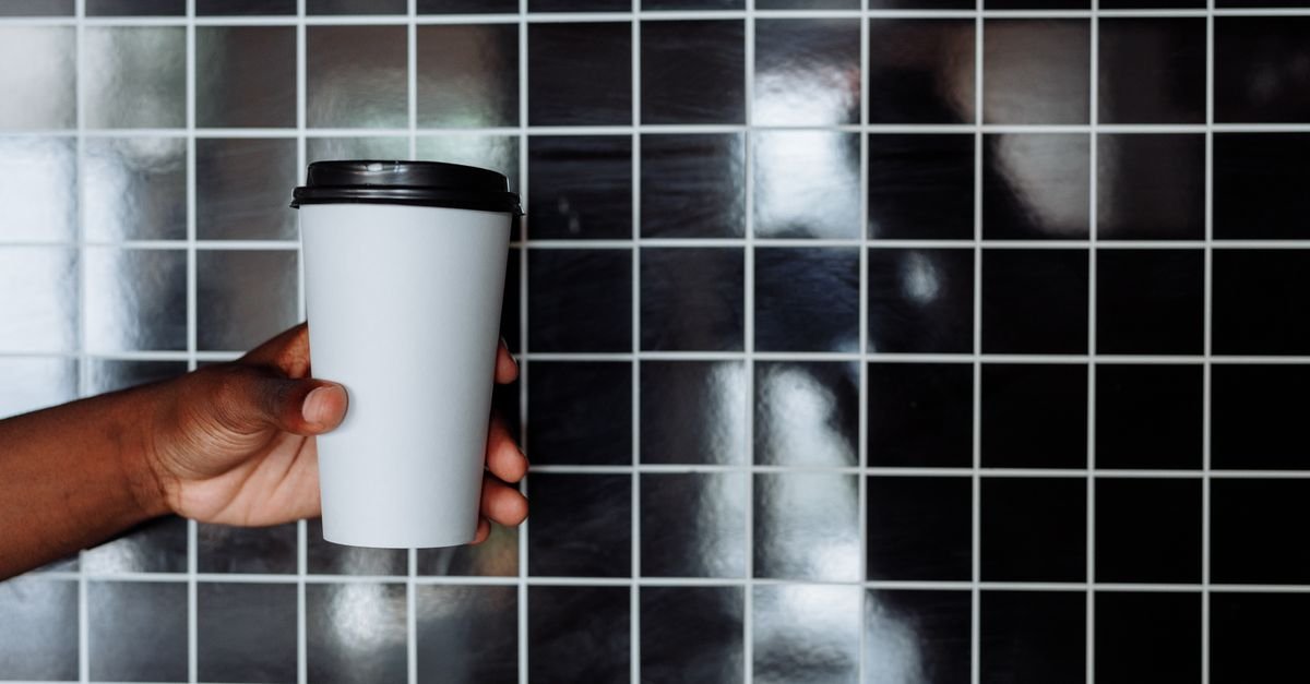 A hand holding a white disposable coffee cup against a tiled black background.