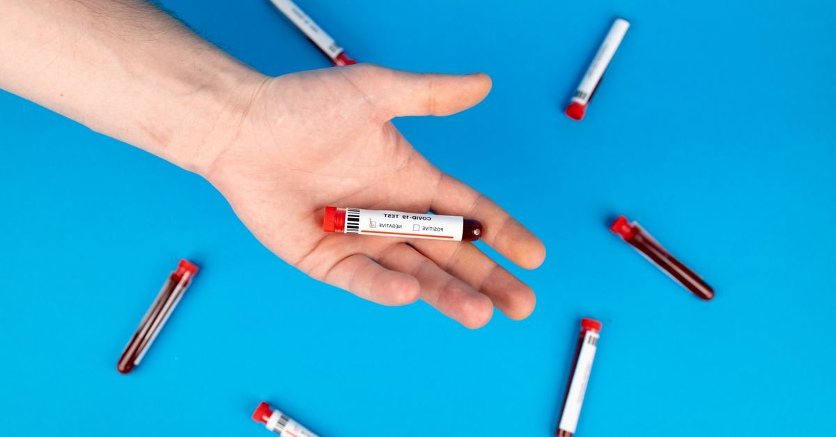Close-up of a hand holding a blood sample vial on blue background, representing COVID-19 testing.