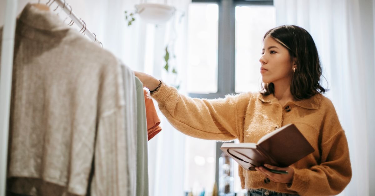 Young Latin American female employee with journal touching assorted wear on stand in house room