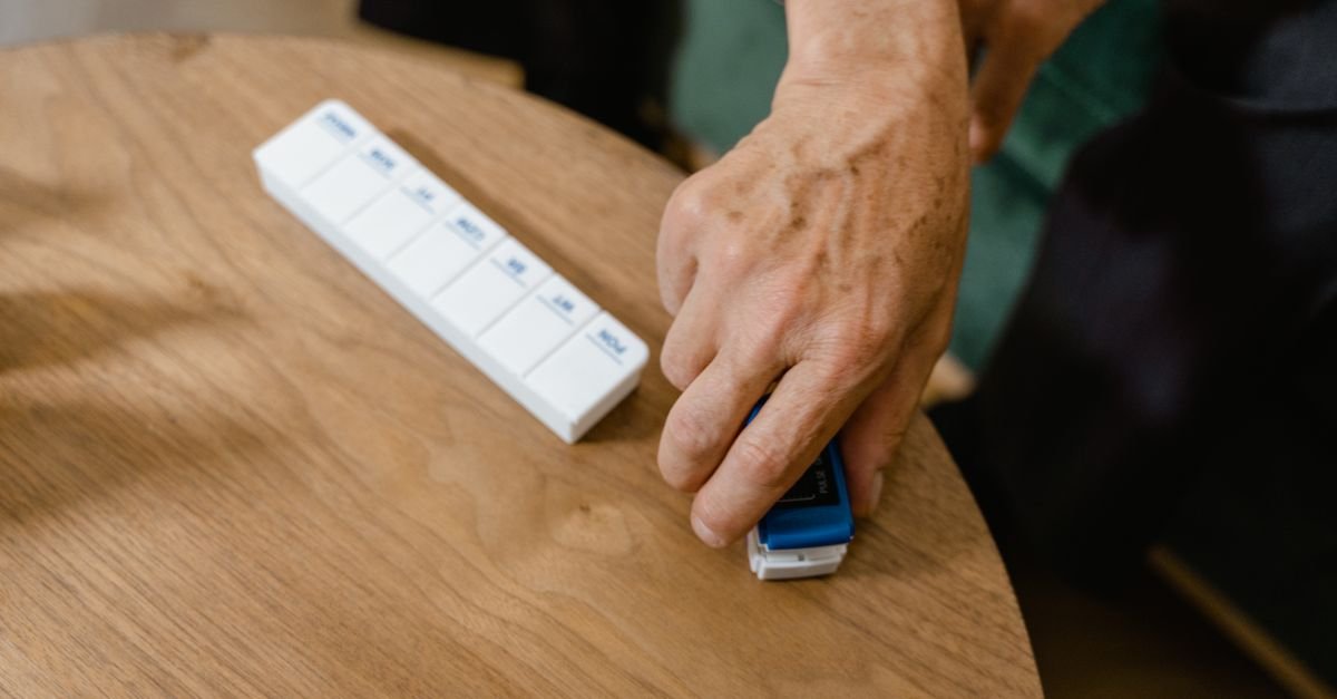 Close-up of an elderly person's hand using a pulse oximeter beside a pill organizer on a table.