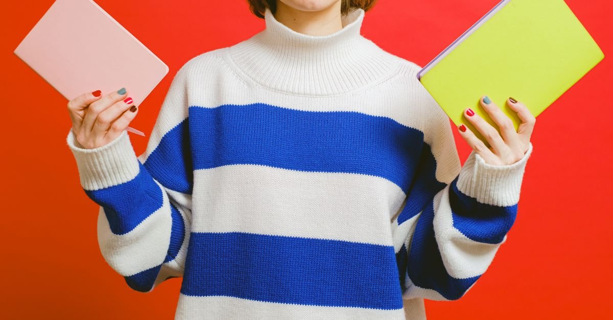 A woman holds pink and yellow notebooks while wearing a white and blue striped sweater against a red background.