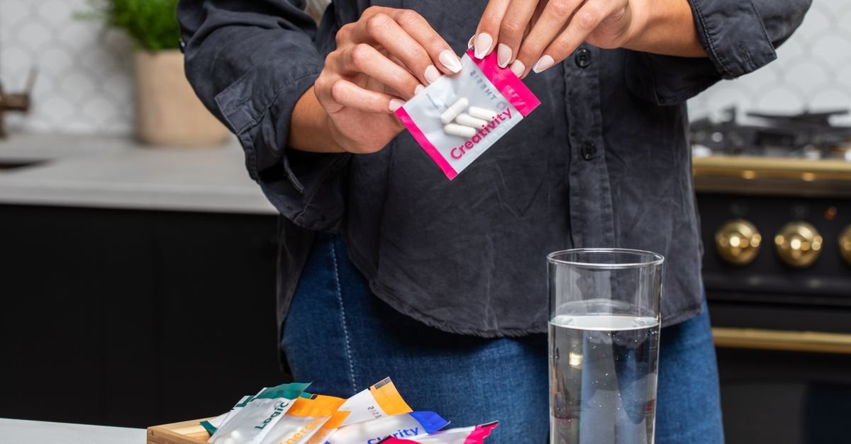 Person opening a creativity supplement pack over a glass of water in a modern kitchen setting.