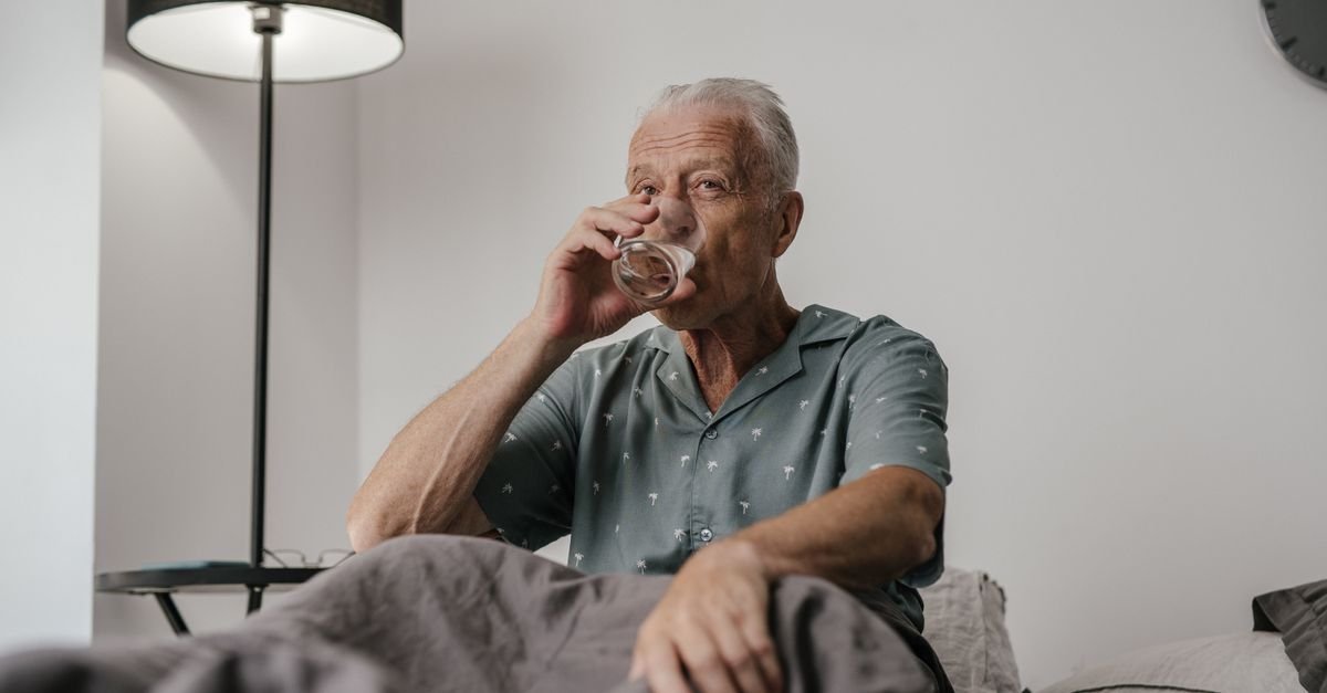 Senior man with gray hair drinks water in bedroom setting, conveying calm and routine.